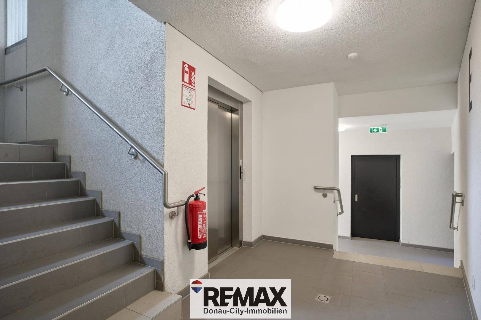 Helles Treppenhaus mit grauen Stufen, Edelstahlgeländer und einem modernen Aufzug.