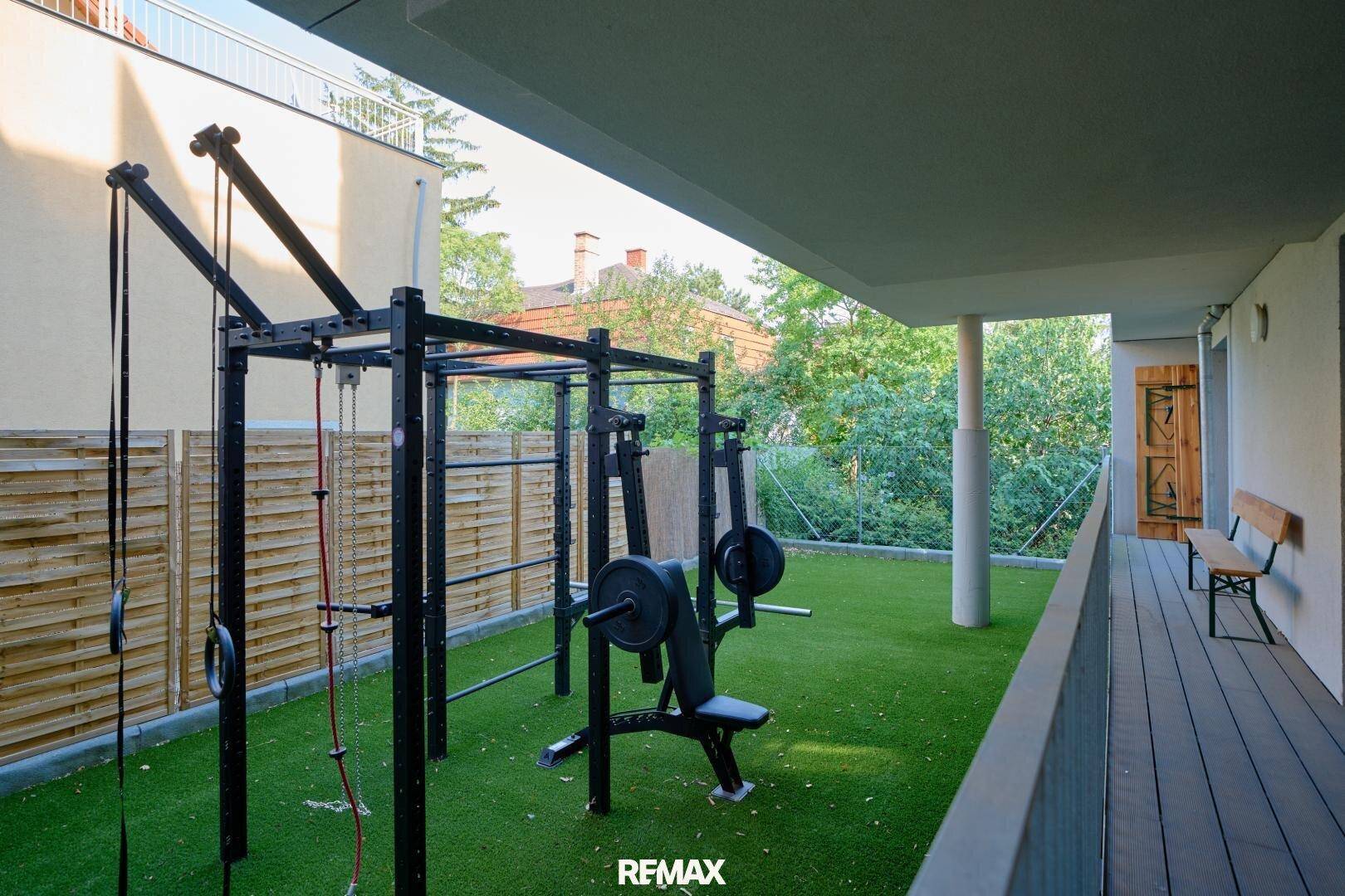 Moderner Außenbereich mit Fitnessgeräten, Kunstrasen und einer Holzterrasse unter einem überdachten Bereich.