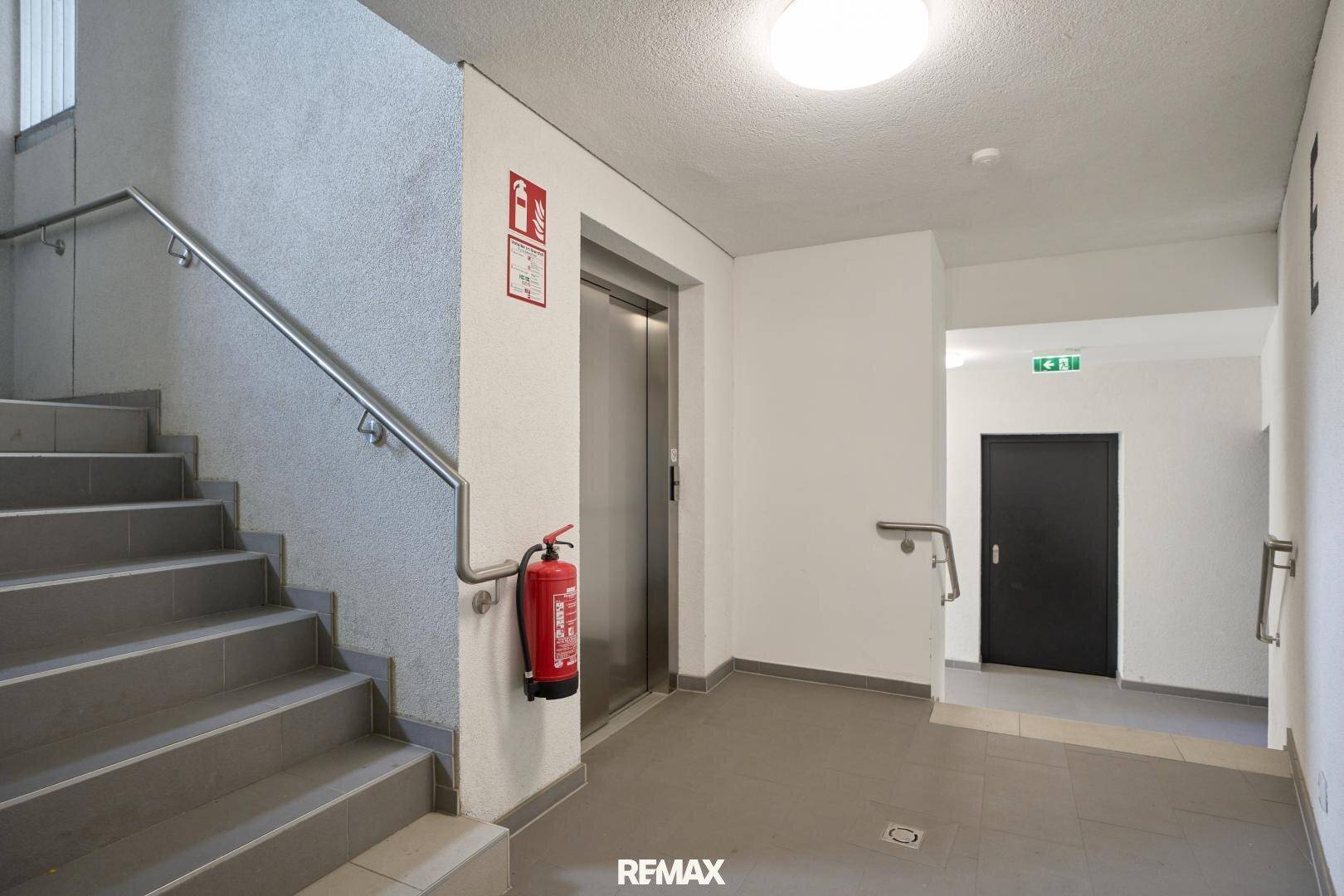 Helles Treppenhaus mit grauen Stufen, Edelstahlgeländer und einem modernen Aufzug für bequemen Zugang.
