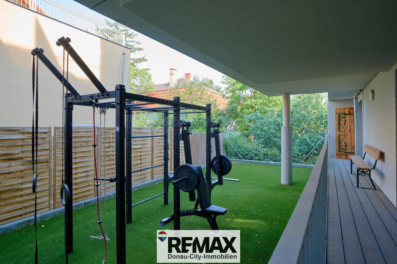 Outdoor-Fitnessbereich mit Trainingsgeräten auf Kunstrasen, umgeben von einem Holzzaun und Bäumen.