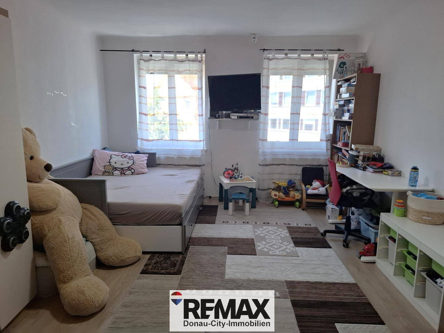 Kinderzimmer mit Bett, Schreibtisch, Spielzeug und zwei Fenstern.