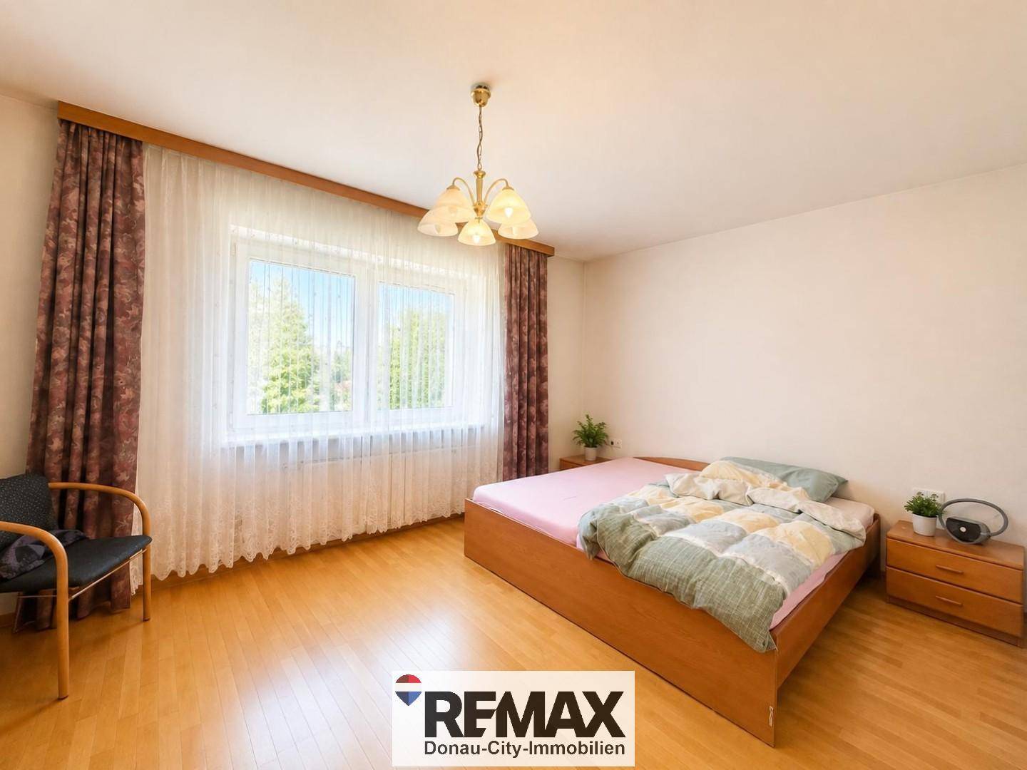 Helles Schlafzimmer mit Parkettboden, Doppelbett und großem Fenster mit Vorhängen.