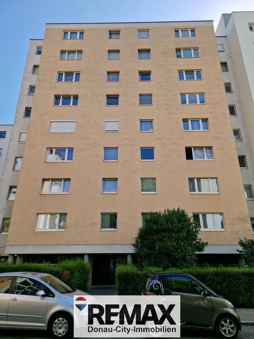 Mehrstöckiges Wohngebäude mit beiger Fassade und zahlreichen Fenstern, umgeben von Grünflächen.