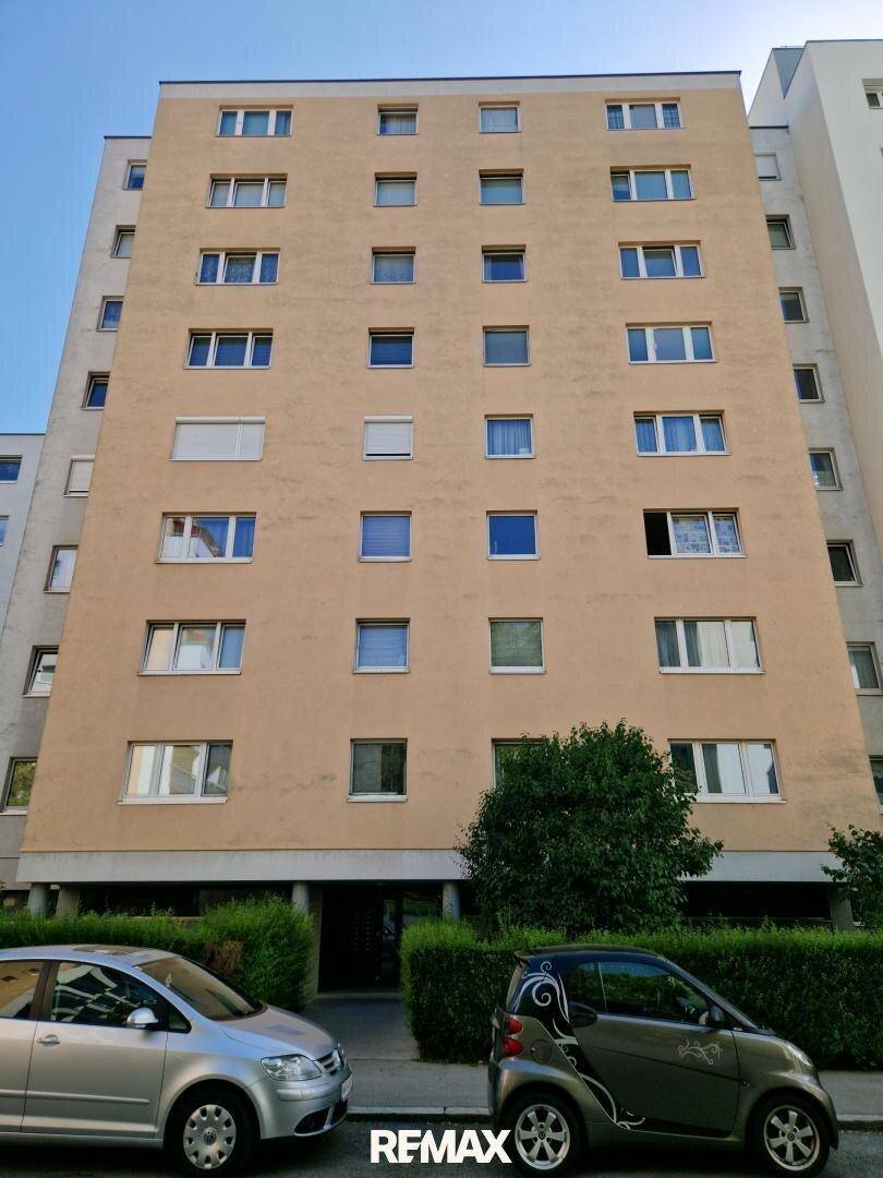 Mehrstöckiges Wohngebäude mit beiger Fassade und zahlreichen Fenstern, umgeben von Grün.