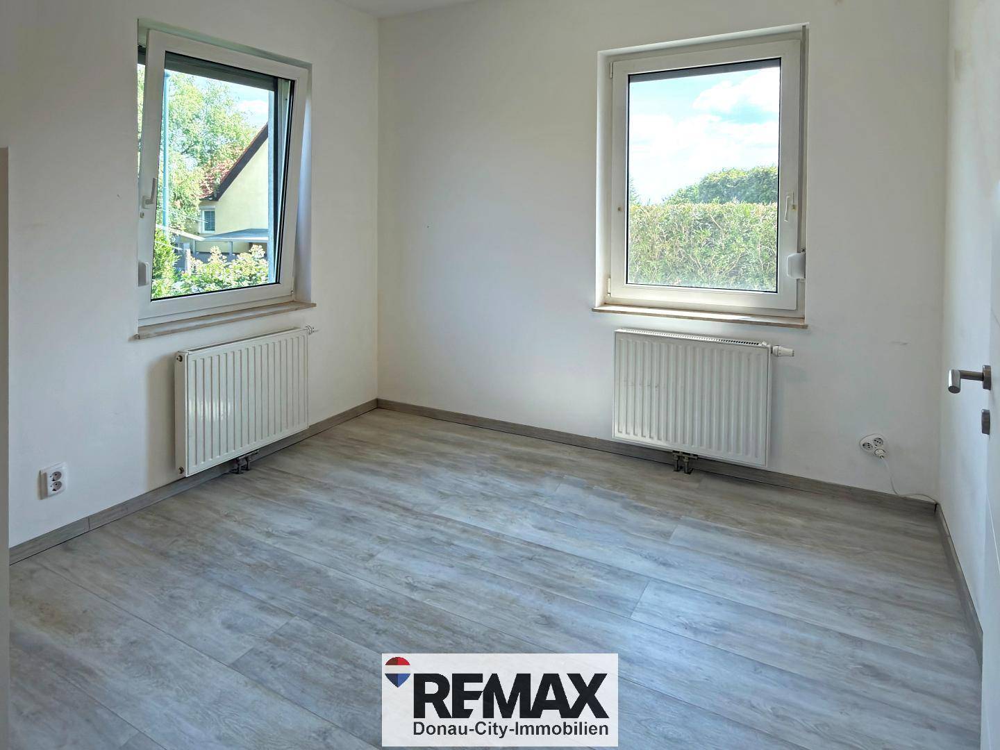 Helles Zimmer mit zwei Fenstern, Heizkörpern und modernem Laminatboden.