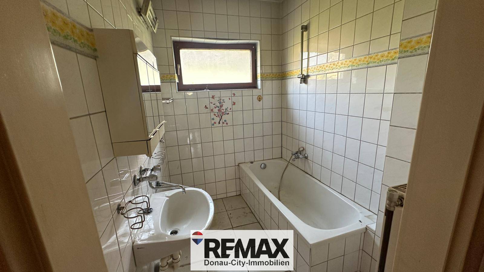 Veraltetes Badezimmer mit weißen Fliesen, Badewanne und kleinem Fenster, renovierungsbedürftig.