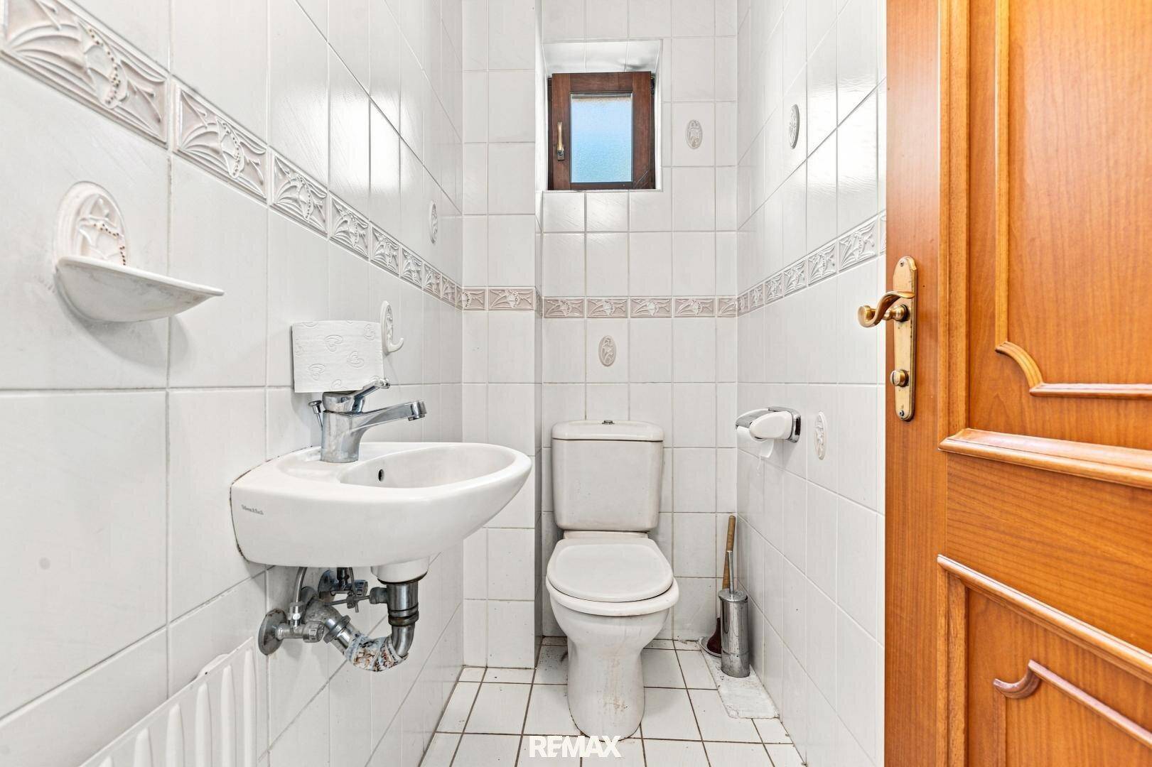 Separate Toilette mit weißen Fliesen, kleinem Waschbecken und Holz-Eingangstür.