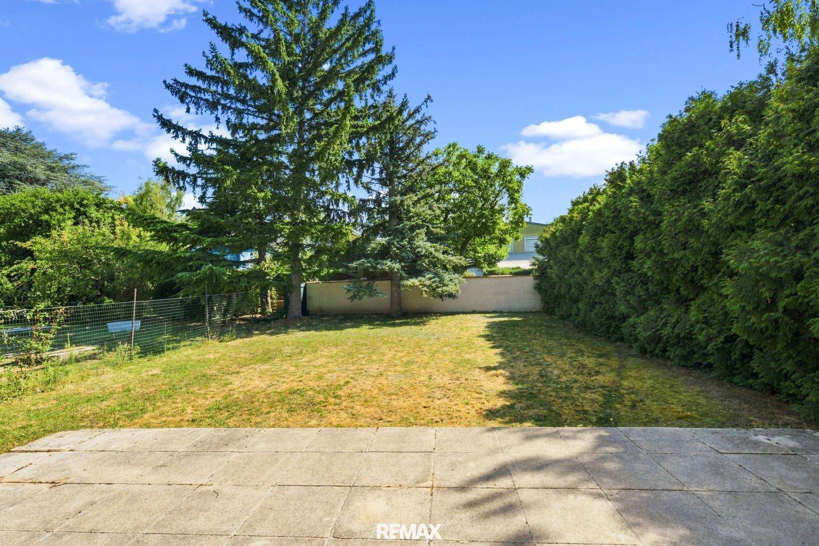 Großer Gartenbereich mit gepflasterter Terrasse und hohen Bäumen, umgeben von Hecken.