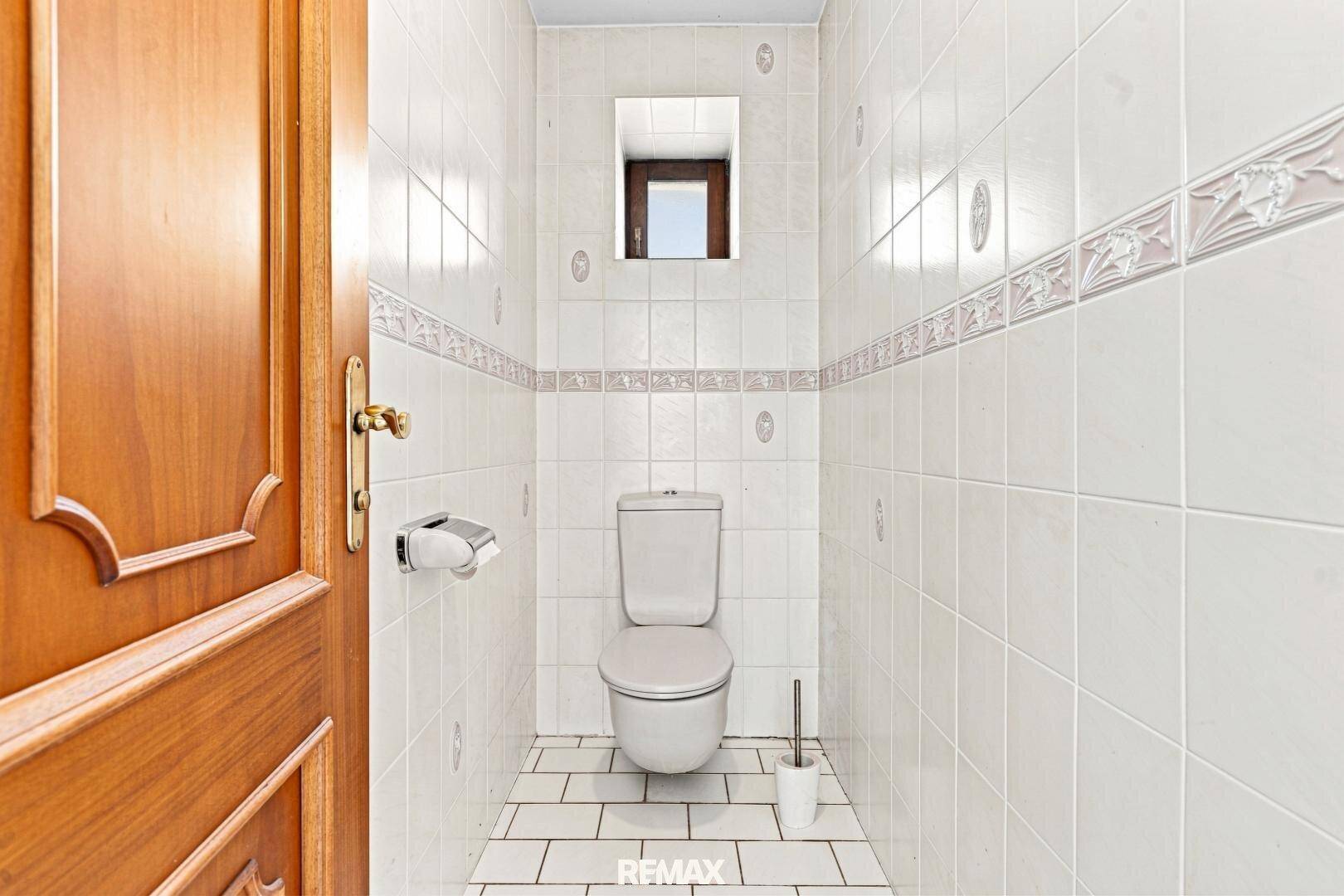 Separate Toilette mit weißen Fliesen, kleinem Fenster und Holz-Eingangstür.