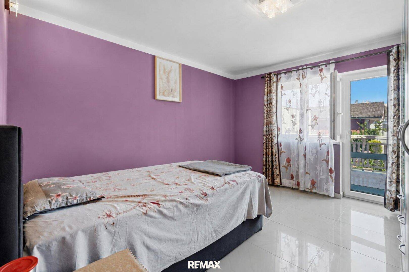 Schlafzimmer mit lila Wänden, Einzelbett und Zugang zu einem kleinen Balkon.