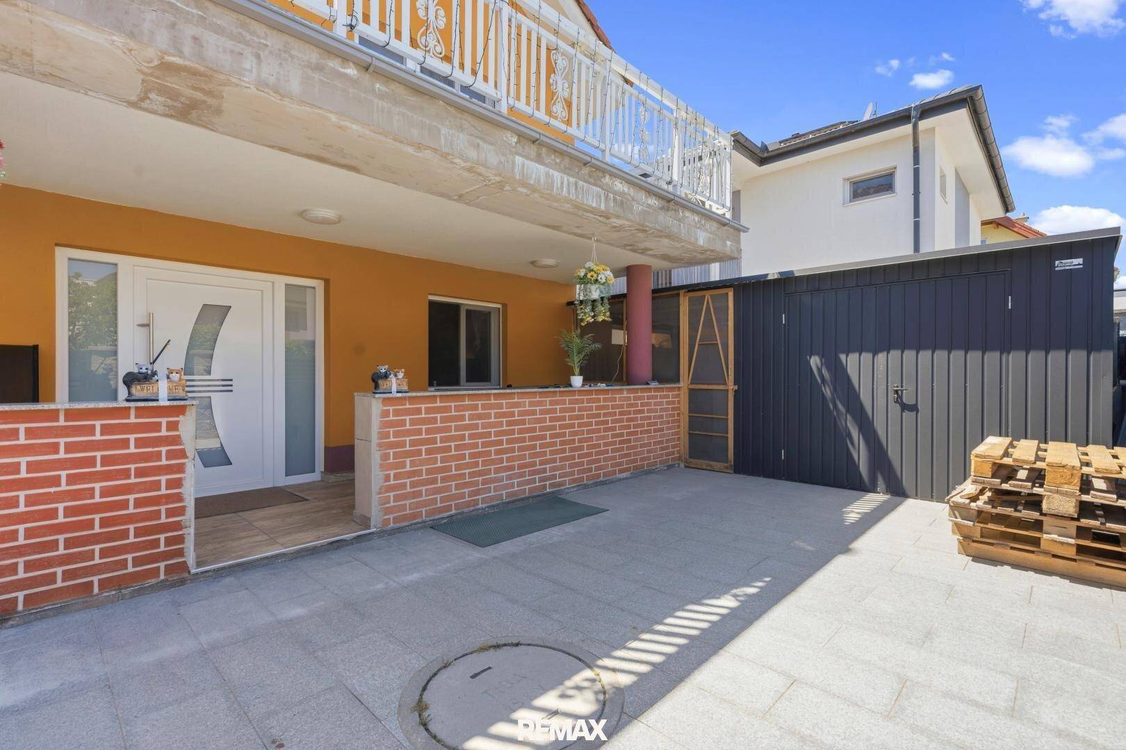 Außenbereich mit gepflasterter Fläche, Eingang und einem Schuppen im Hintergrund.