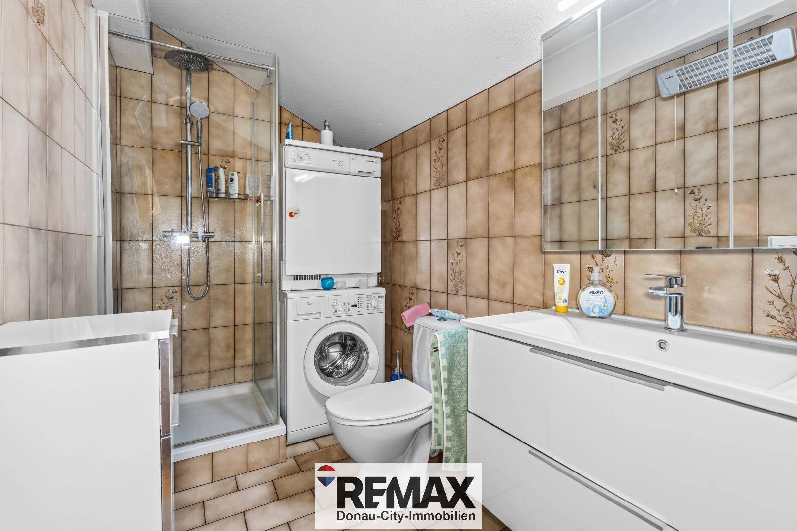Badezimmer mit Dusche, WC, Waschmaschine und Trockner, gefliest in warmen Brauntönen.