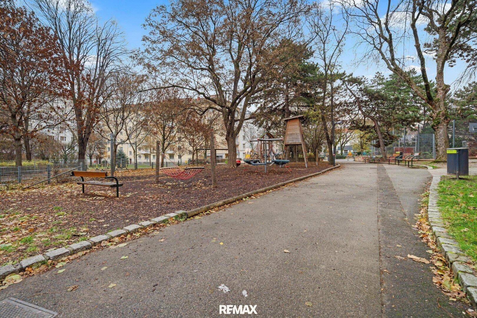 Großer öffentlicher Spielplatz mit Bäumen und verschiedenen Spielgeräten in einer städtischen Umgebung.