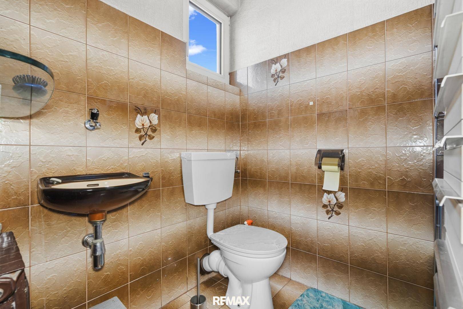 Separate Toilette mit braunen Fliesen, kleinem Waschbecken und einem Fenster für Tageslicht.