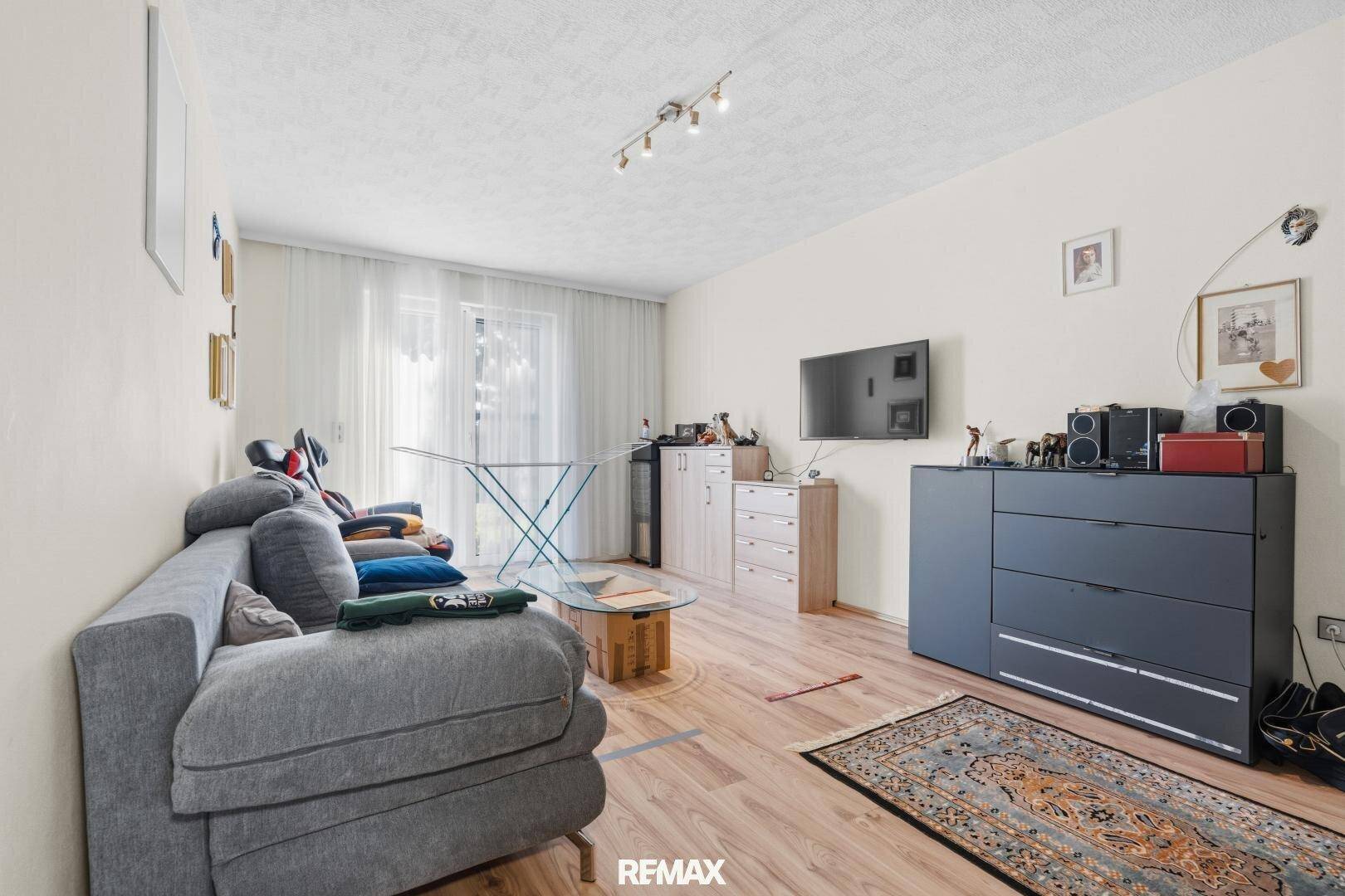 Gemütliches Wohnzimmer mit hellem Laminatboden, bequemer Couch und Blick ins Grüne durch das Fenster.
