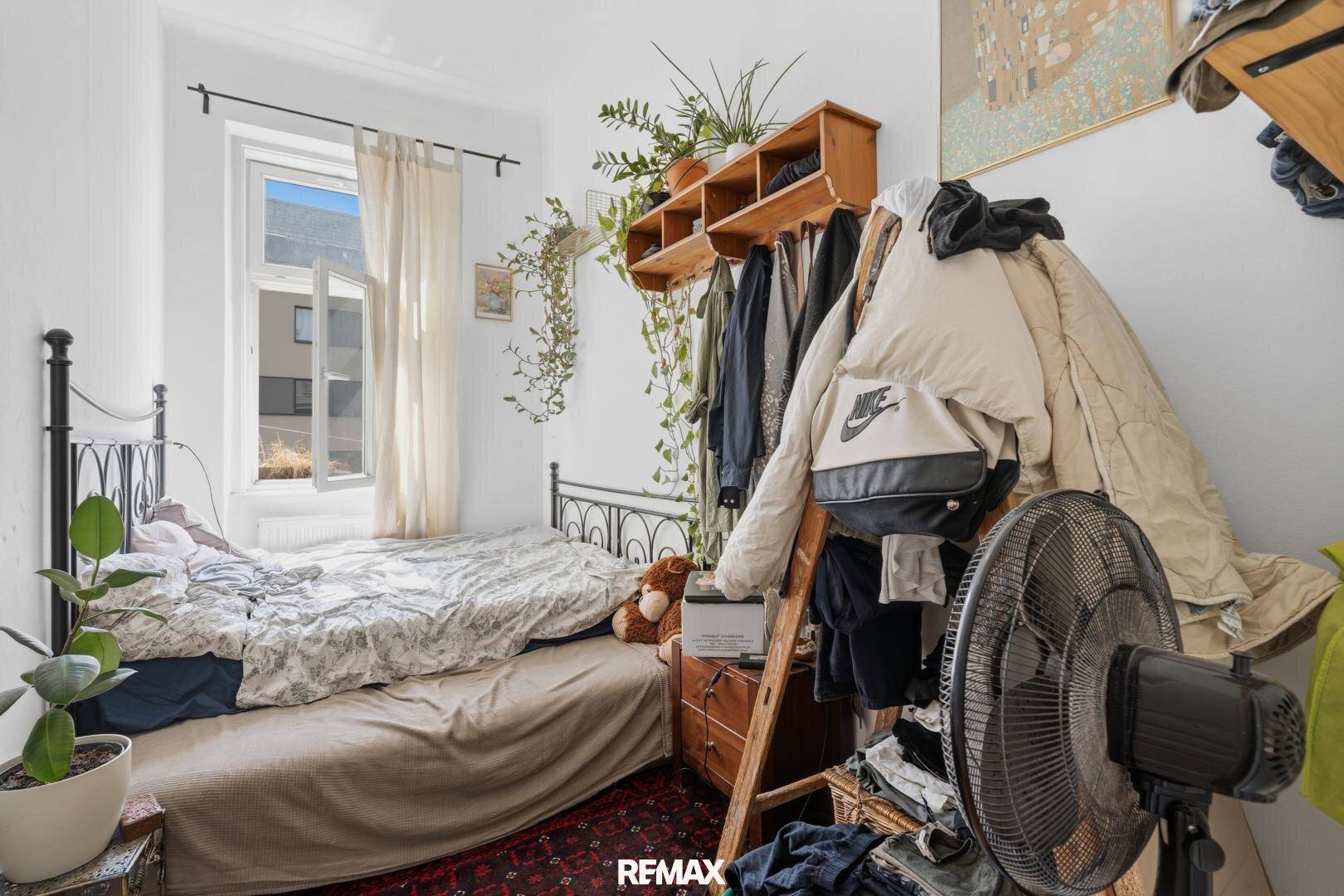Schlafzimmer mit Bett, Kleiderstange und Fenster, das viel Licht hereinlässt und eine angenehme Helligkeit bietet.