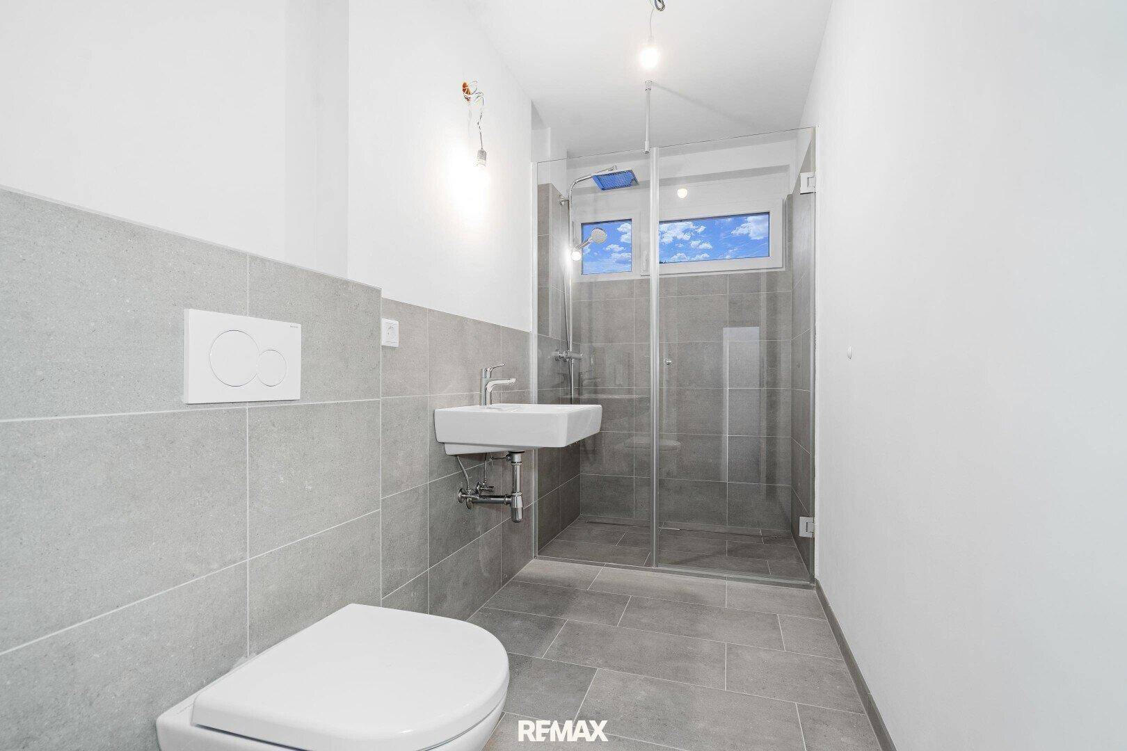 Modernes Badezimmer mit Dusche, Waschbecken und WC, gefliest in hellen Grautönen.