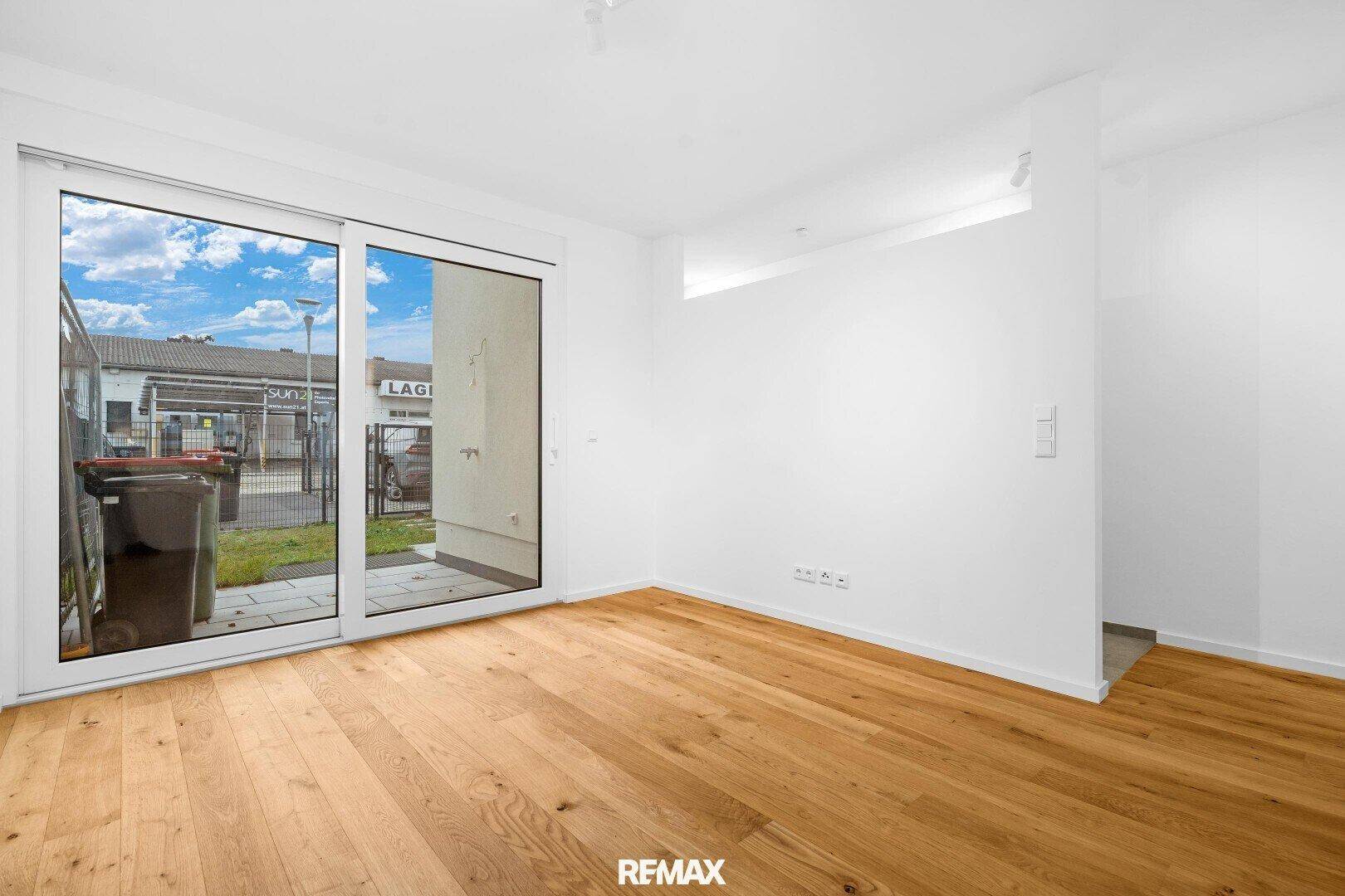 Heller, leerer Raum mit Holzboden und großem Fenster mit Blick auf den Garten.