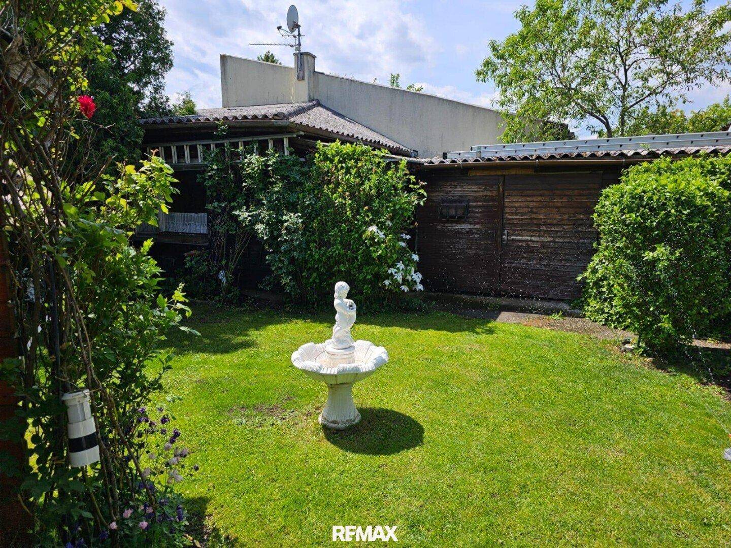Sonniger Garten mit Rasenfläche, einem dekorativen Brunnen und einem Holzschuppen im Hintergrund.
