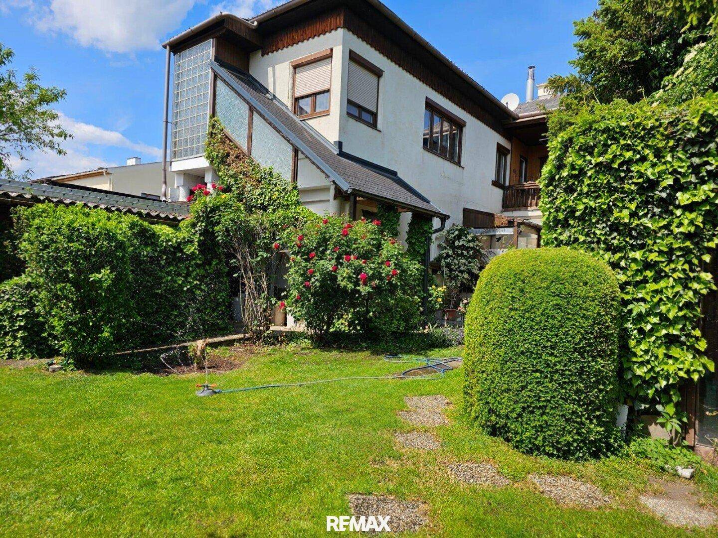 Rückansicht des Hauses mit üppigem Garten, hohen Hecken und viel Grünfläche unter blauem Himmel.