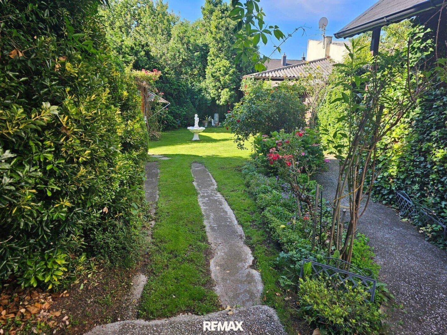 Sonniger Garten mit Rasenfläche, Gartenweg und blühenden Pflanzen entlang des Hauses.