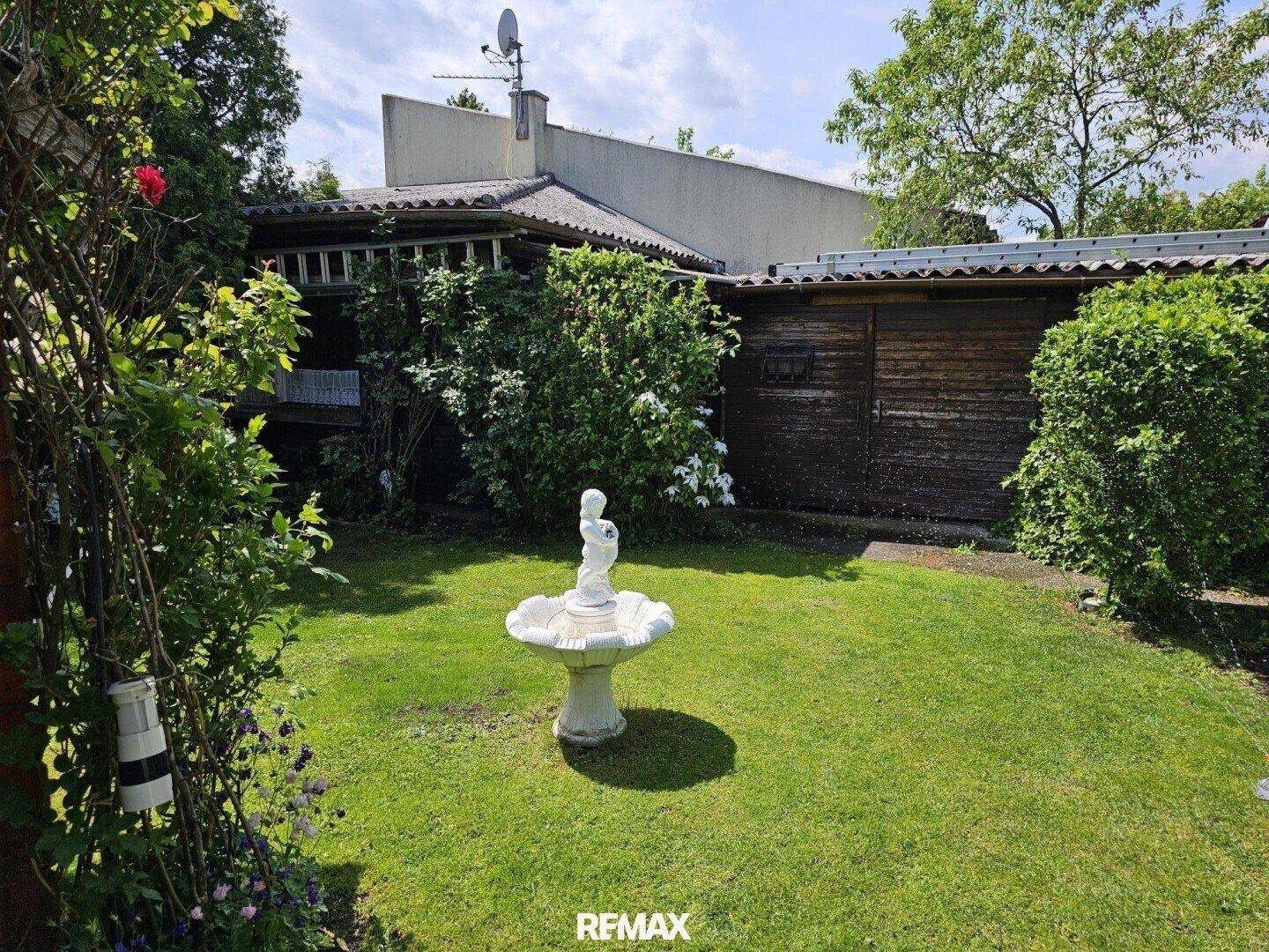 Sonniger Garten mit Rasenfläche, einem dekorativen Brunnen und einem Holzschuppen im Hintergrund.