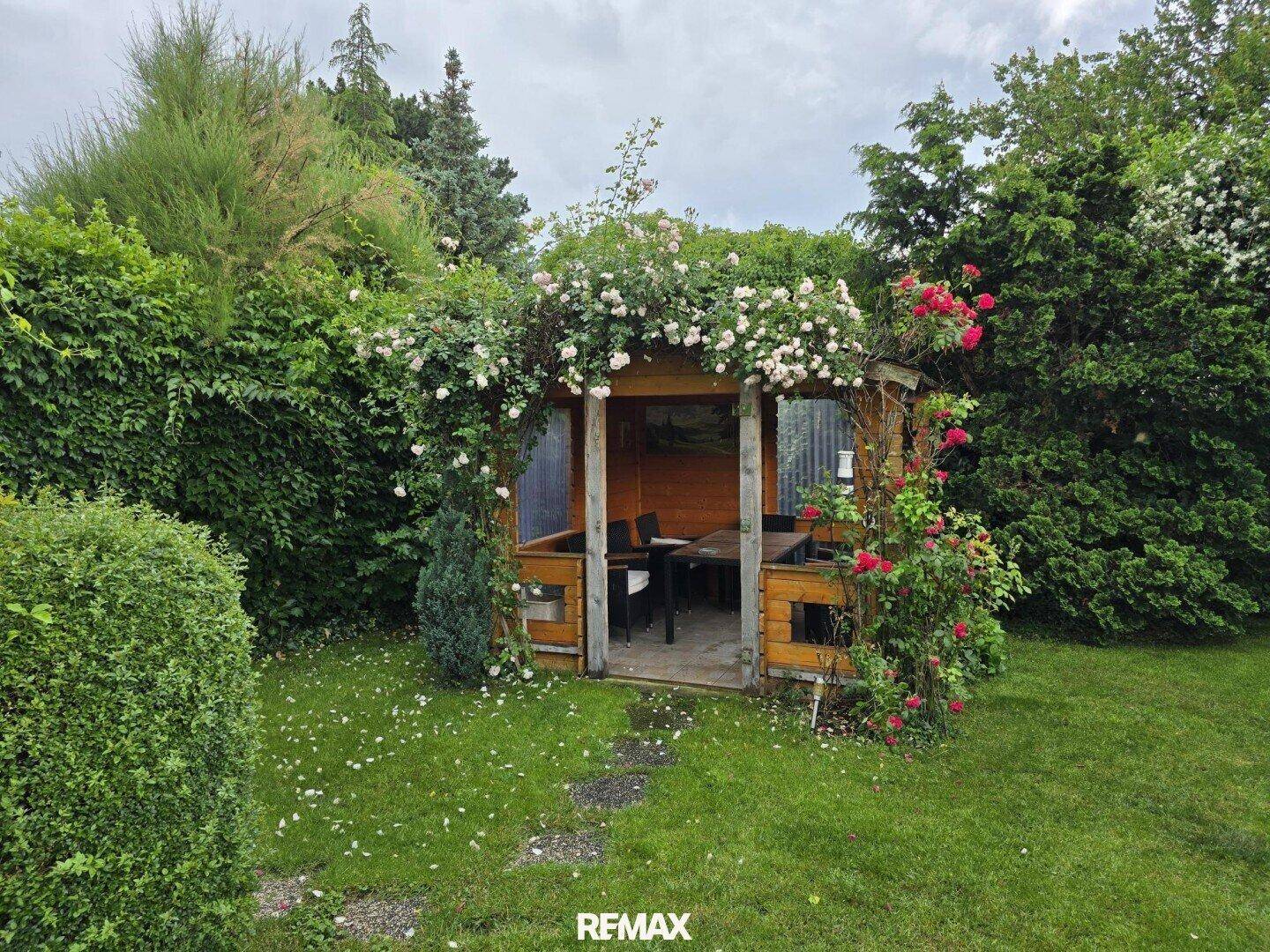 Charmantes Gartenhaus mit Sitzgelegenheiten, umgeben von blühenden Rosen und grünen Büschen.