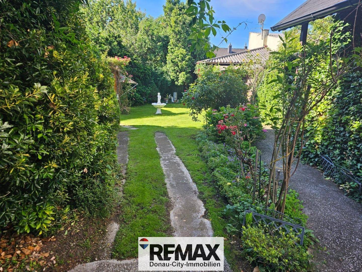 Sonniger Garten mit Rasenfläche, Wegen, einem Brunnen und üppiger Bepflanzung.