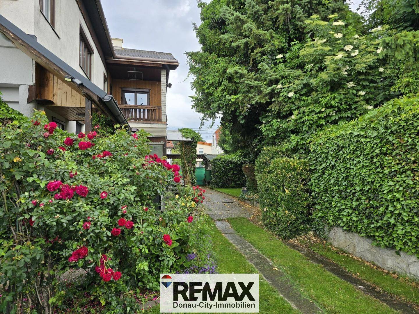 Gartenweg führt entlang des Hauses, umgeben von üppigen Rosenbüschen und hohen Bäumen.
