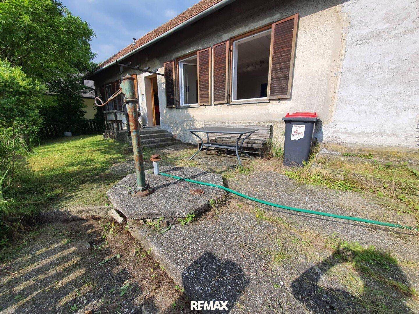 Seitenansicht eines Hauses mit einer alten Wasserpumpe und einem kleinen gepflasterten Bereich.