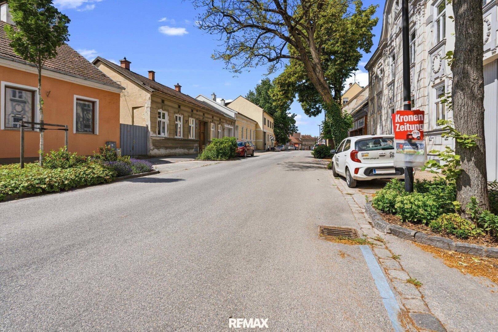 Sonnige Straßenansicht mit alten Gebäuden, Bäumen und parkenden Autos unter blauem Himmel.