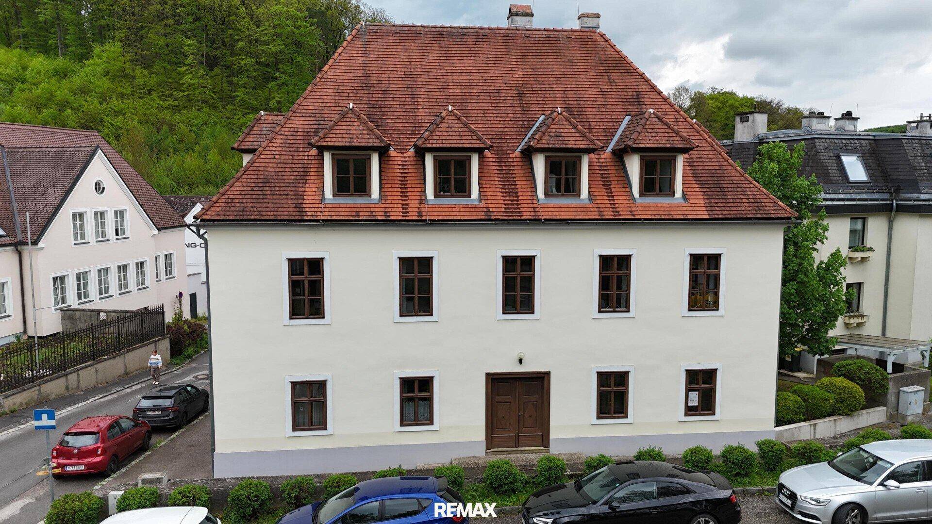 Historisches Gebäude mit rotem Ziegeldach und mehreren Gaubenfenstern in ländlicher Umgebung.
