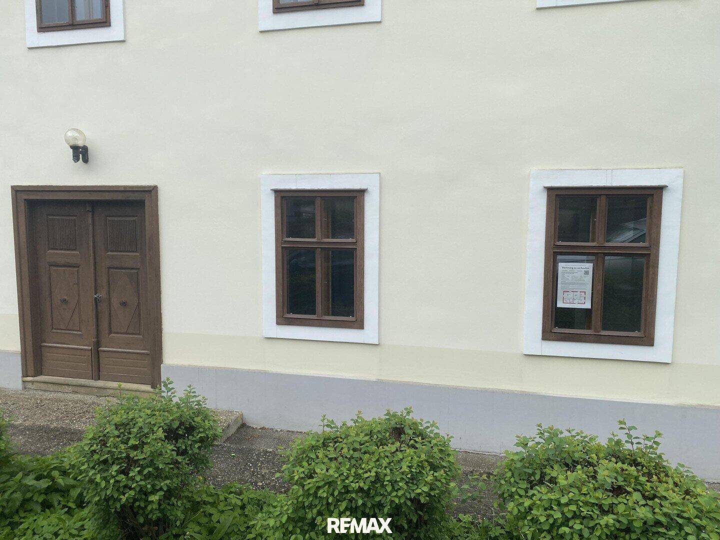 Detailansicht der Fassade mit Holztür und Fenstern, umgeben von grünen Büschen.