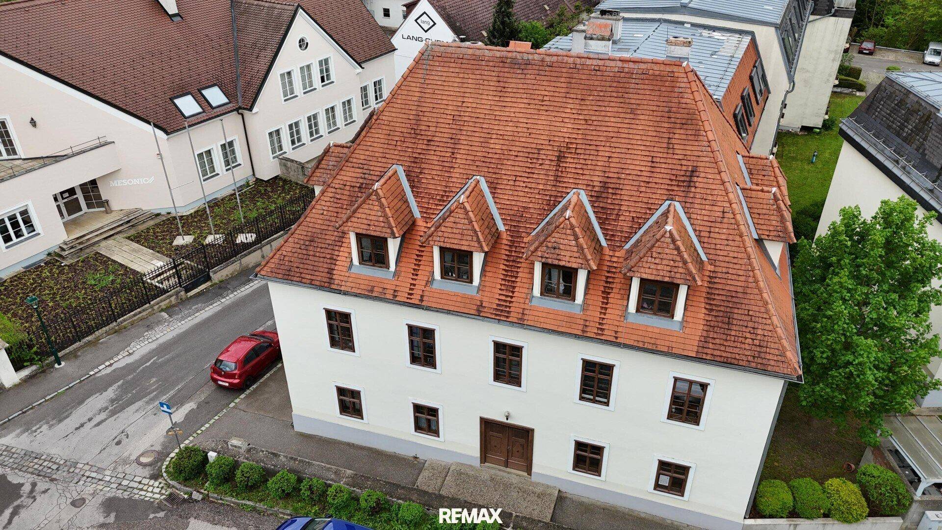 Historisches Gebäude mit rotem Ziegeldach und Gaubenfenstern, umgeben von weiteren Häusern.