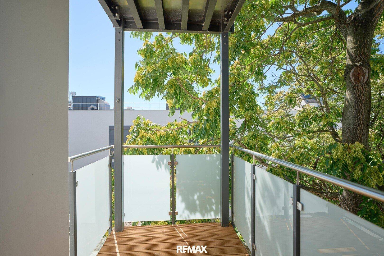 Sonniger Balkon mit Holzbelag und Glasgeländer, umgeben von grünen Bäumen.