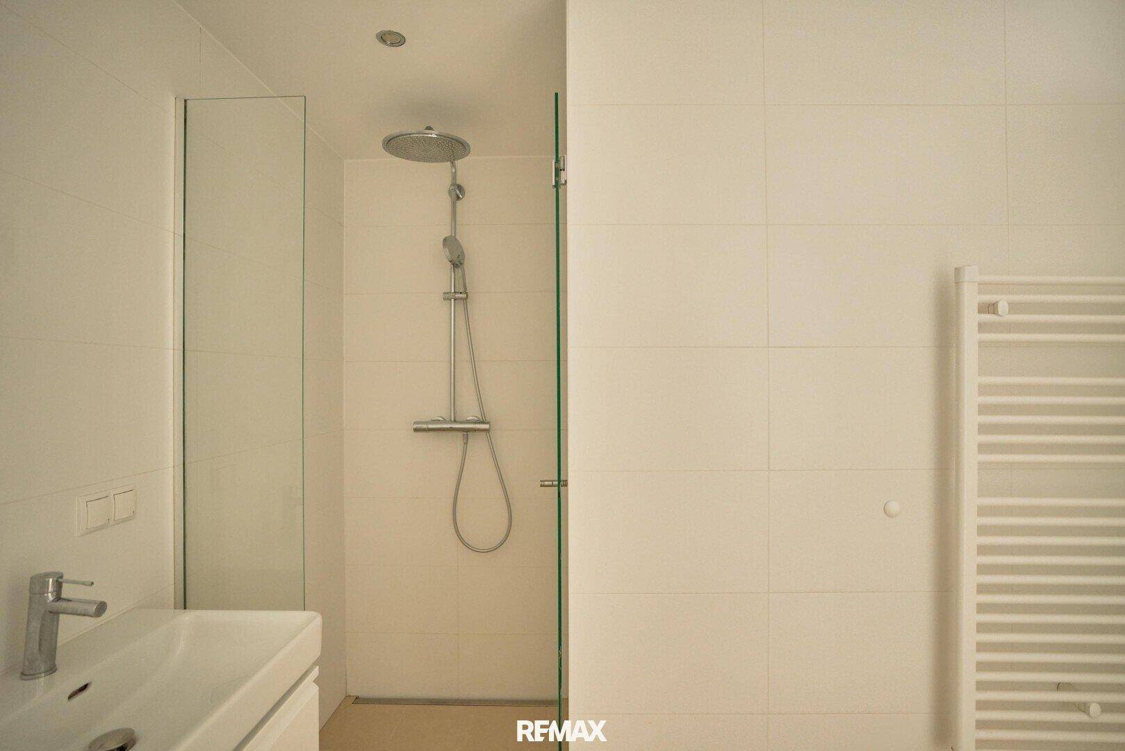 Helles Badezimmer mit weißen Fliesen, begehbarer Dusche und Regenduschkopf.