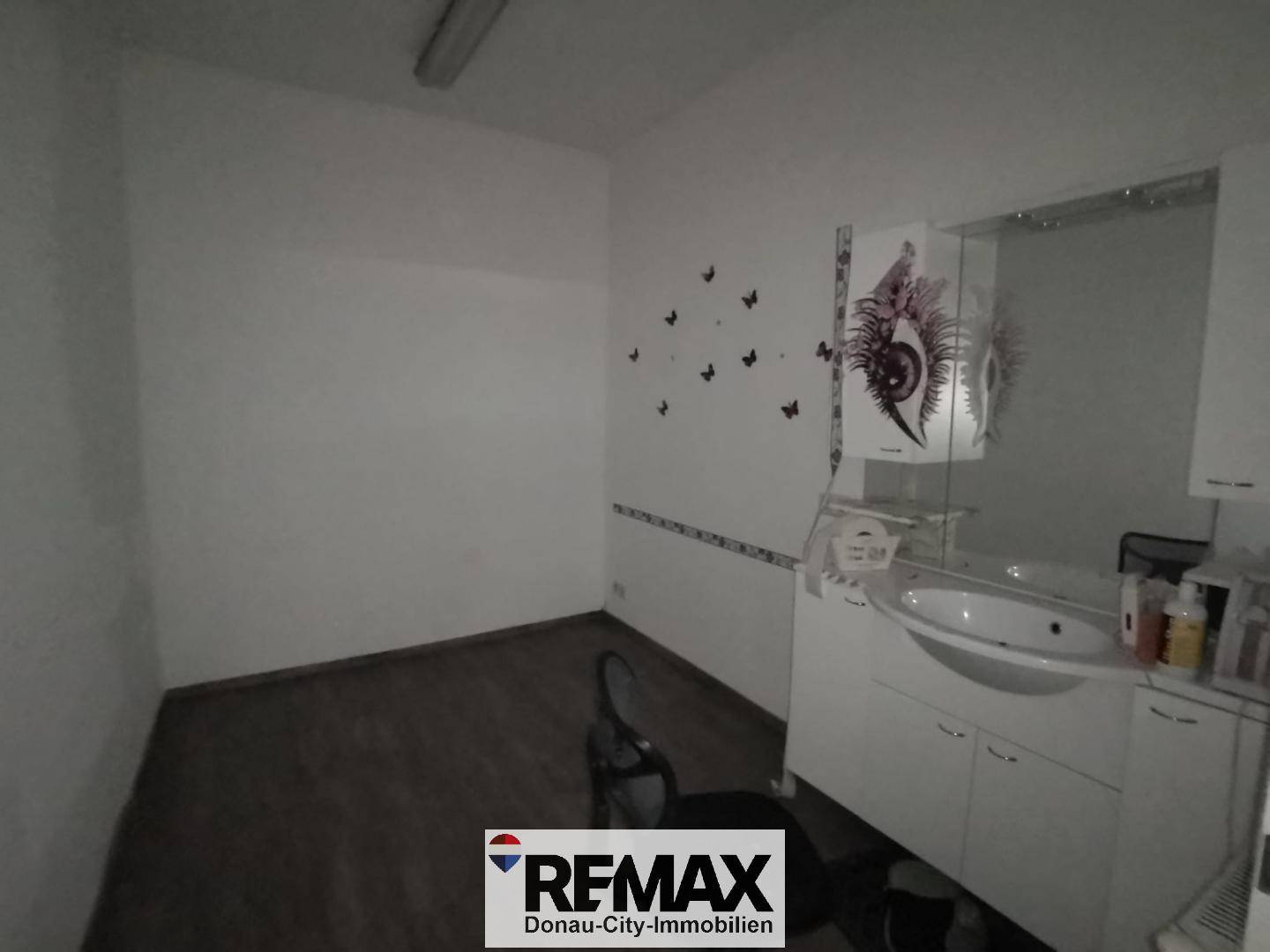 Kleiner Behandlungsraum mit Waschbecken, Spiegel und Wanddekoration in dunklerer Beleuchtung.