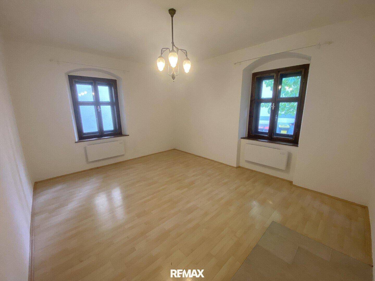 Helles, leeres Zimmer mit Laminatboden, weißen Wänden und zwei Fenstern, ausgestattet mit einem Kronleuchter.