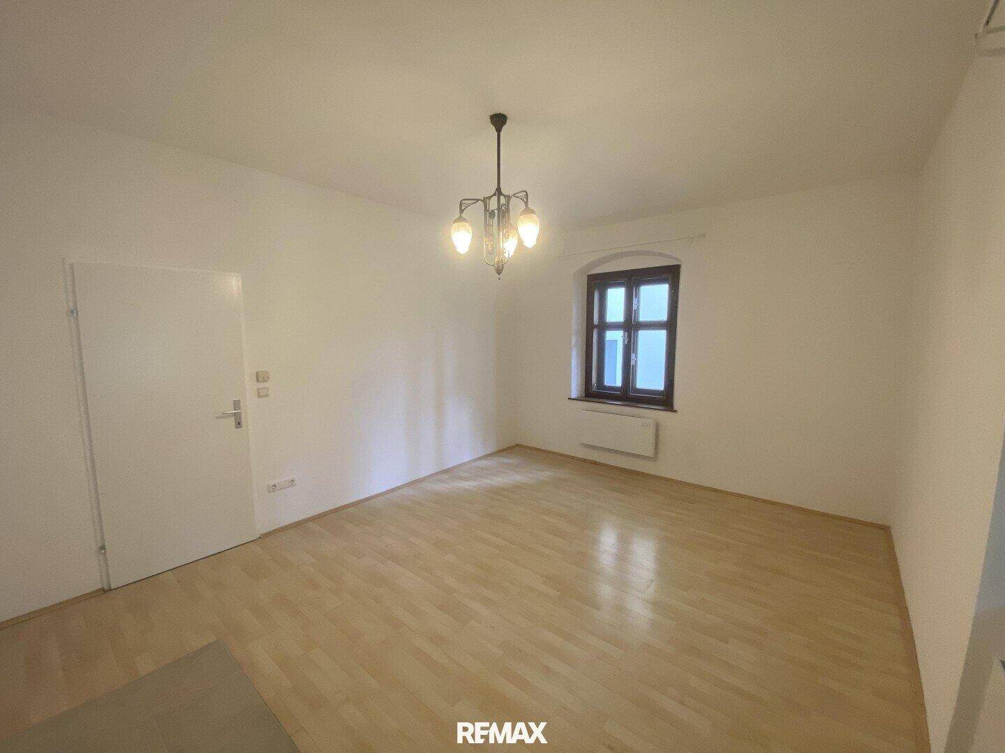 Helles Zimmer mit Laminatboden, weißen Wänden und einem Fenster, beleuchtet von einem klassischen Kronleuchter.