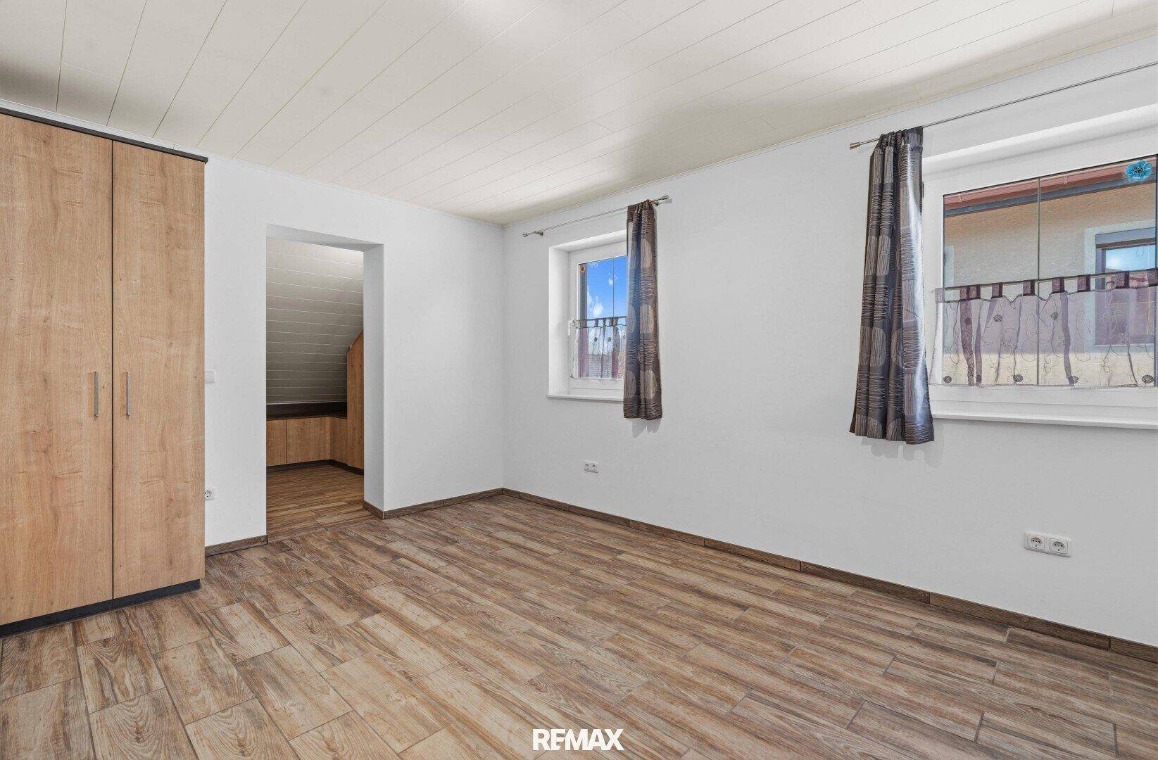 Helles Schlafzimmer mit Laminatboden, Holzdecke und zwei Fenstern.