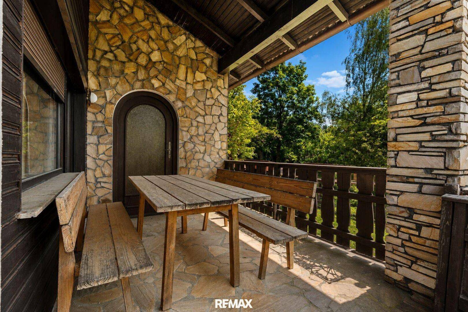 Überdachter Balkon mit rustikalen Steinwänden, Holztisch und Bänken, umgeben von viel Grün.