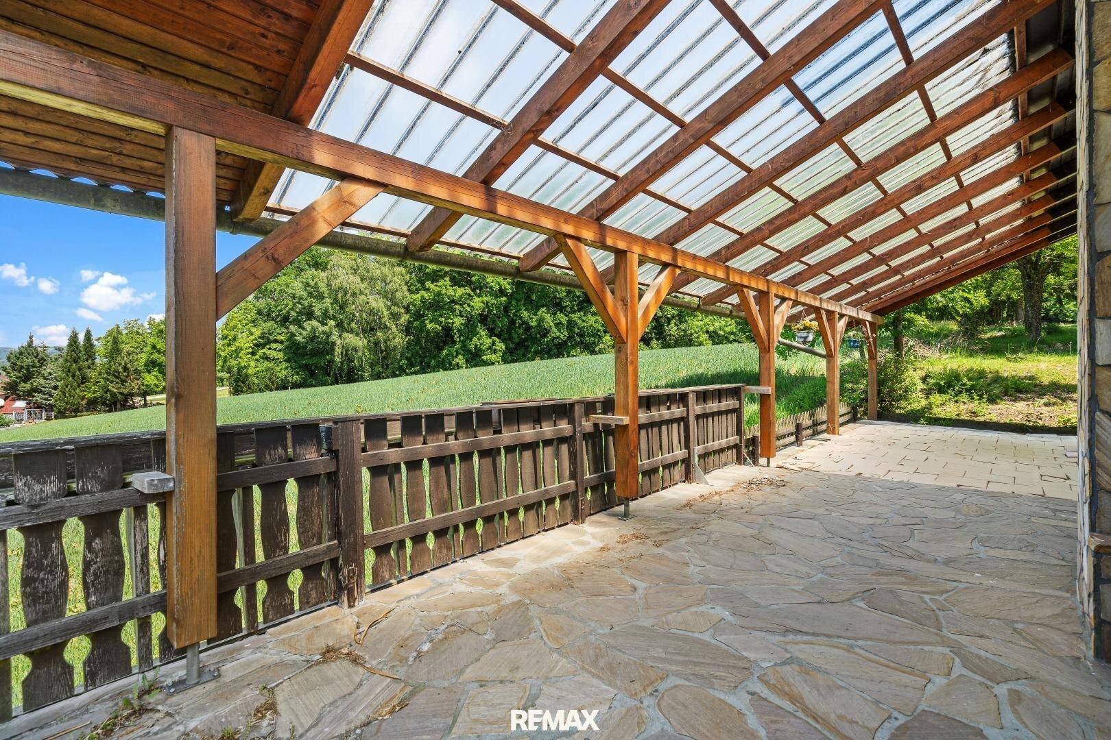 Überdachte Terrasse mit Steinboden und Holzbalken, umgeben von Natur.