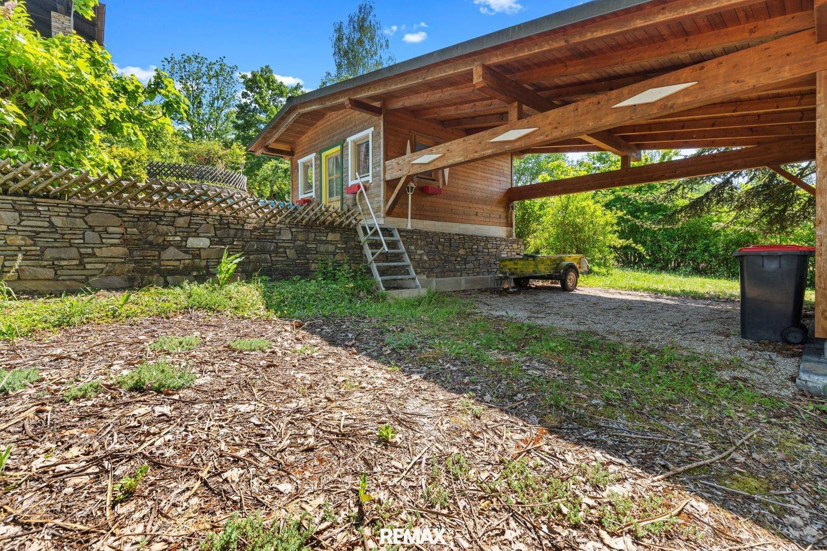 Überdachter Carport aus Holz neben einem kleinen Nebengebäude im Grünen.