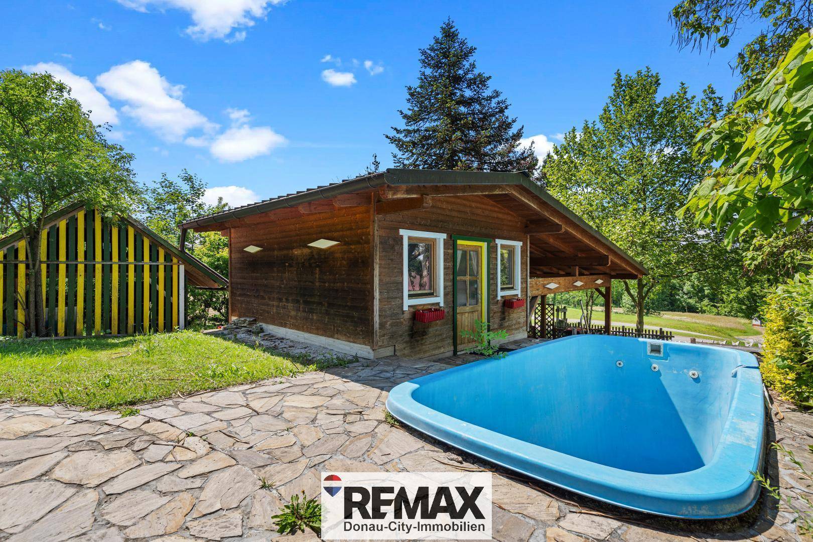 Kleiner Pool in einem gepflasterten Gartenbereich neben einem Holzhaus unter blauem Himmel.