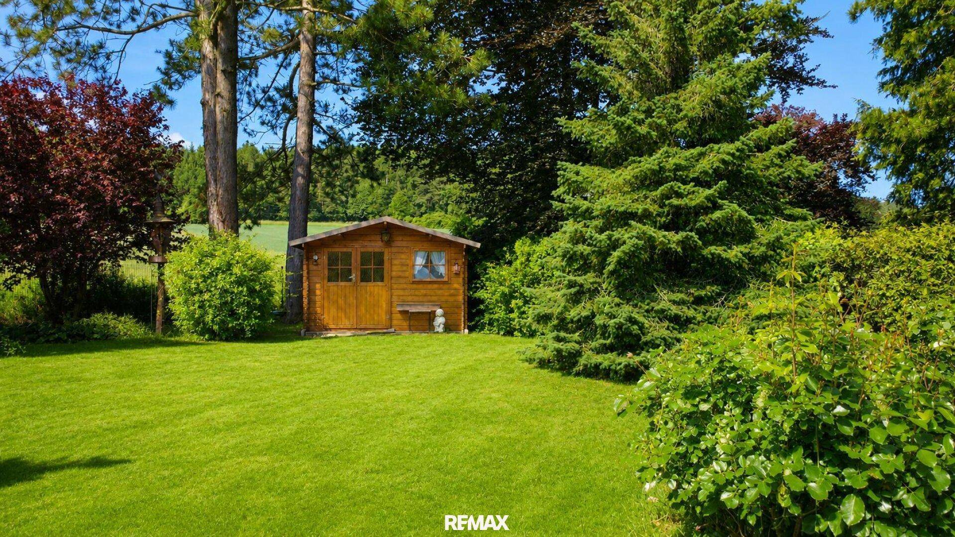 Großer, gepflegter Garten mit Rasenfläche und einem kleinen Holzgartenhaus unter hohen Bäumen.