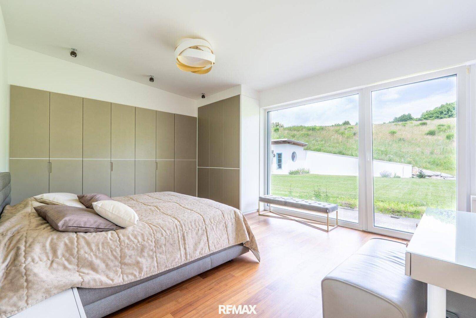 Schlafzimmer mit Holzboden, Einbauschränken und großen Fenstern mit Gartenblick.