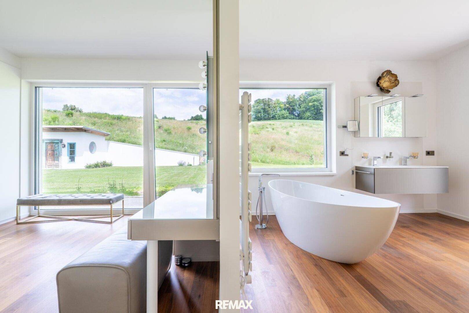 Helles Badezimmer mit freistehender Badewanne, Waschtisch und weitem Blick ins Grüne.