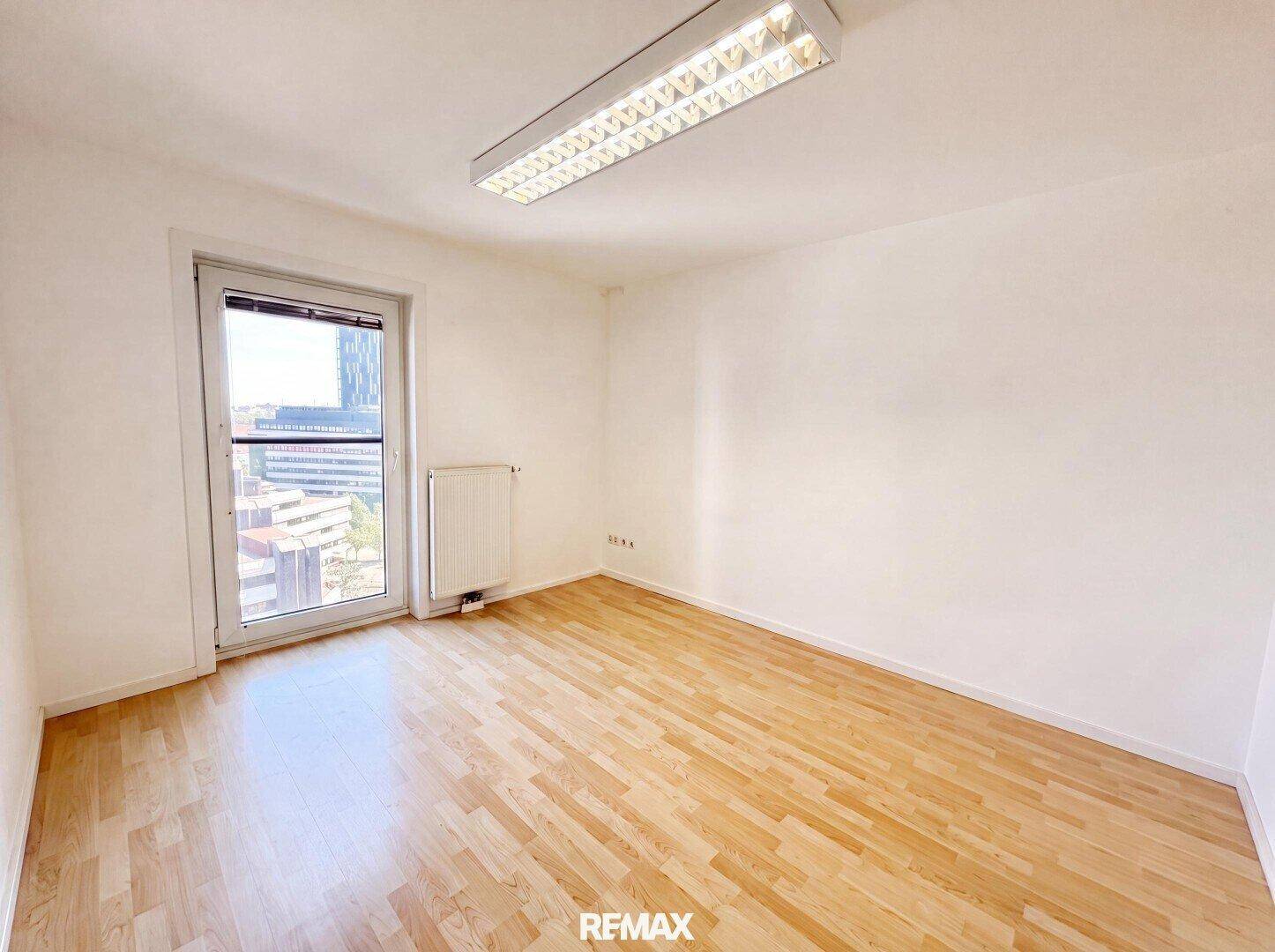 Leeres Schlafzimmer mit hellem Holzboden, weißen Wänden und einem Fenster mit Stadtblick.