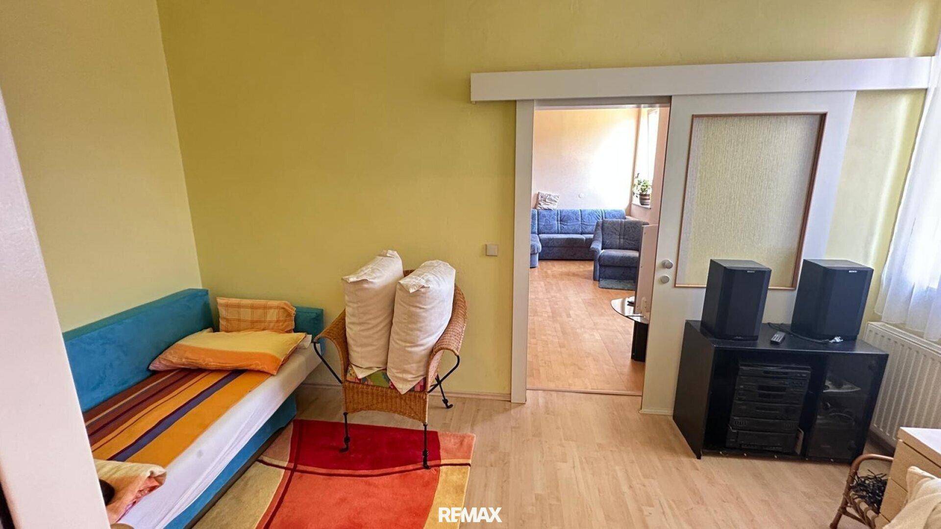 Helles Zimmer mit Tagesbett und Schiebetür, die zu einem weiteren Wohnbereich führt.