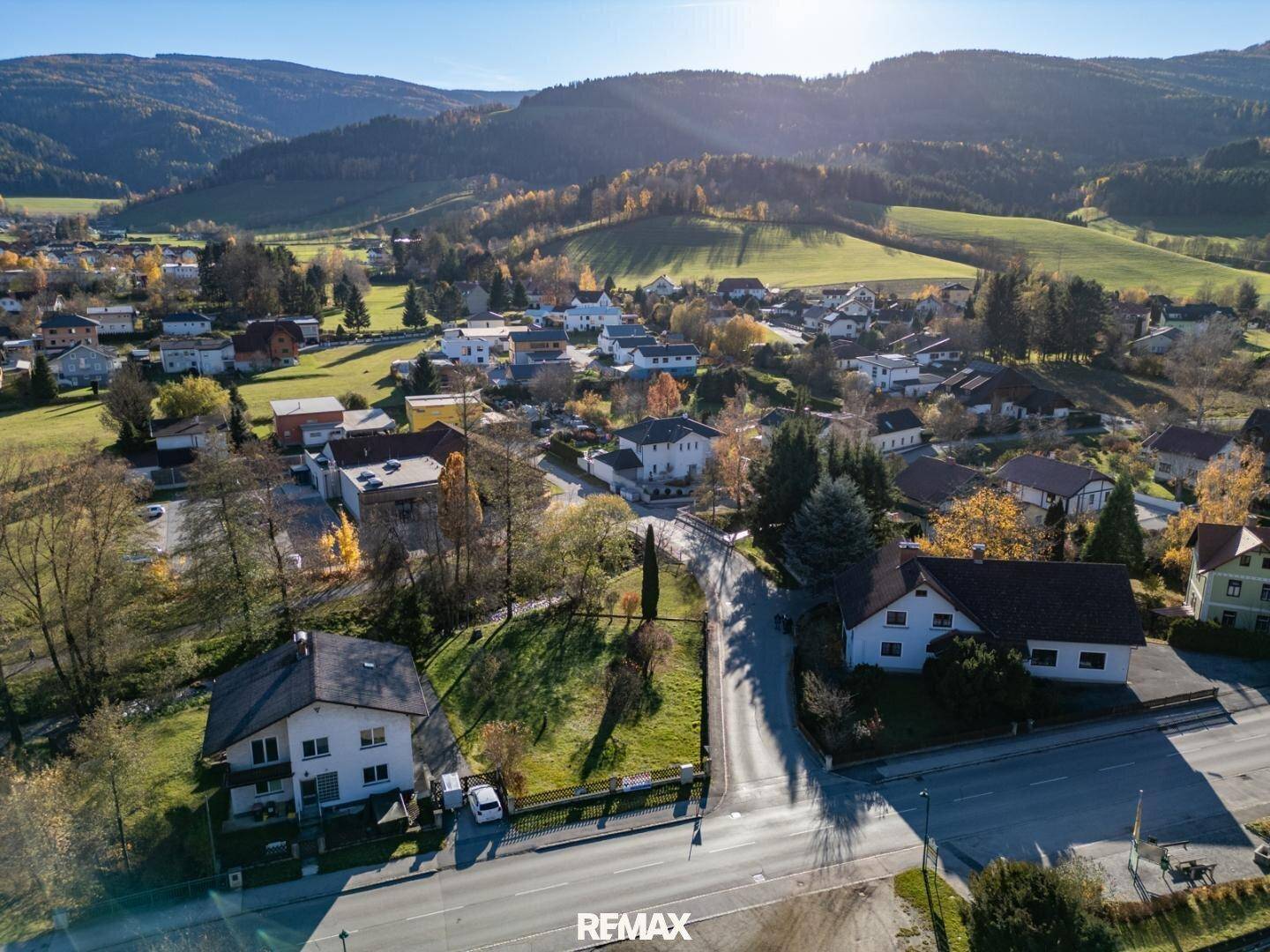 Weitläufige Luftaufnahme einer ländlichen Siedlung, eingebettet in eine malerische Hügellandschaft unter blauem Himmel.